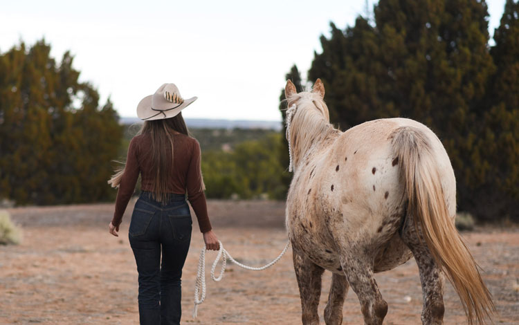 Una mujer, Stella Maria Baer, con un sombrero de vaquero color canela y conduciendo un caballo appaloosa lejos de la cámara hacia los árboles.